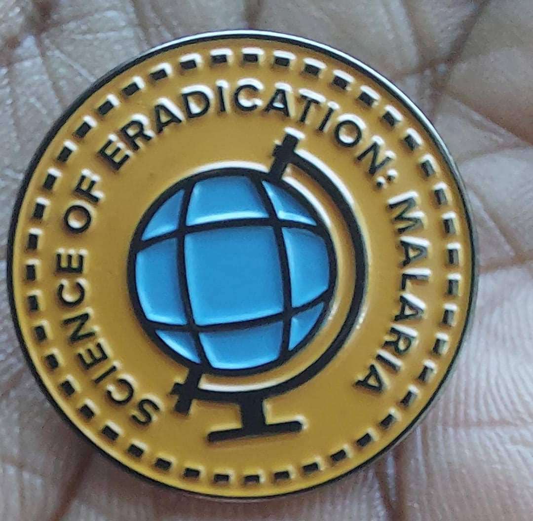 An alum holding a small metal lapel pin of "Science of Eradication: Malaria" in the palm of their hands