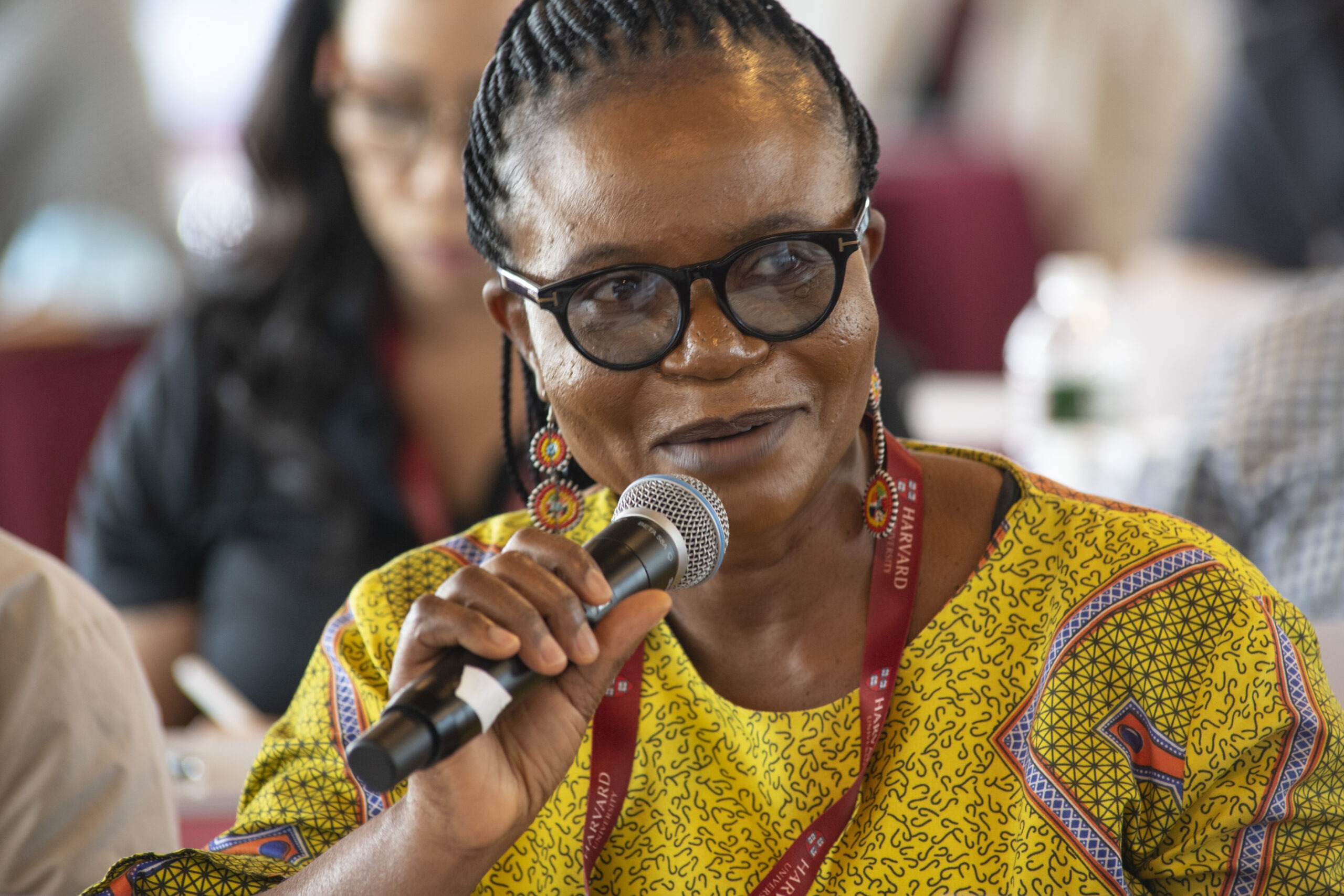 Lorretta Favour Ntoimo speaks into a microphone, wearing a colorful patterned dress. The setting is the "Science of Eradication: Malaria" at the Smith Center in Boston, MA, USA in 2022.